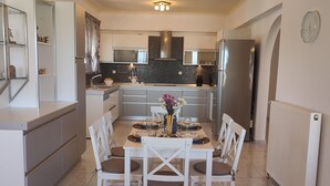 A modern kitchen with a dining area, featuring a central island, white cabinetry, and a tiled backsplash. - Aithrea's Homes in Pastida Rhodes (Pastida)