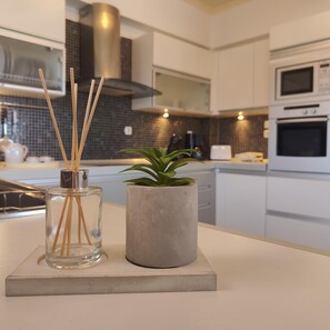 Una cocina moderna con armarios blancos, una campana extractora de acero inoxidable y un microondas. Hay un florero de cristal transparente con palitos de madera y una planta en maceta sobre la encimera.