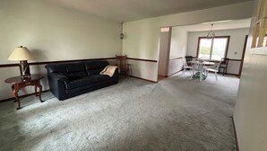 Uma sala de estar com um sofá preto, uma mesa de madeira pequena com um abajur, e um espaço de jantar com uma mesa e cadeiras.