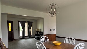 Uma área de refeições com uma mesa de madeira e cadeiras brancas, um candelabro e vista para uma sala de estar com um sofá e uma luminária.