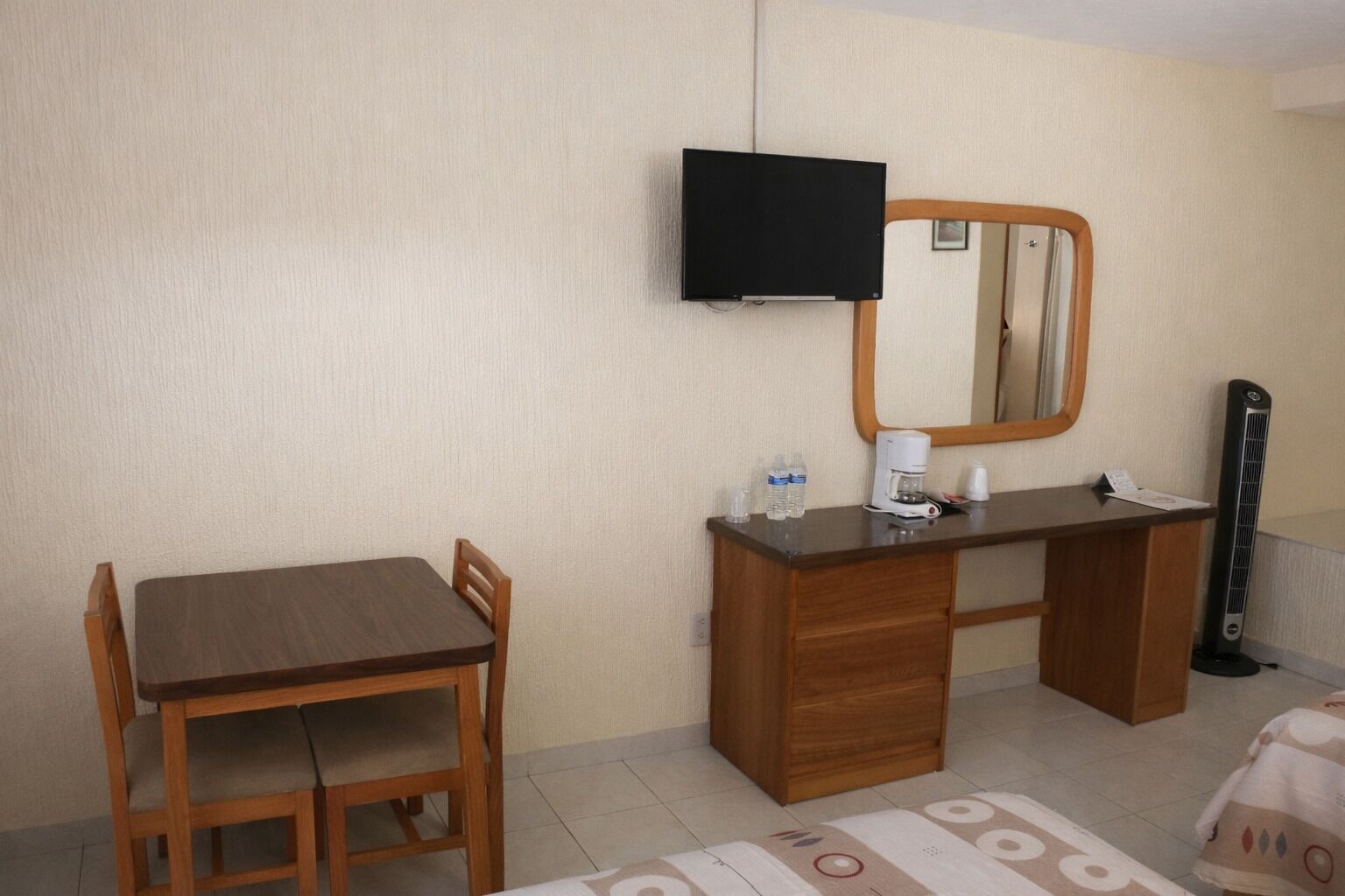 Habitación de hotel con un escritorio de madera, una silla, televisor montado en la pared, un espejo, un calentador y una mesita con una cafetera y botellas de agua.