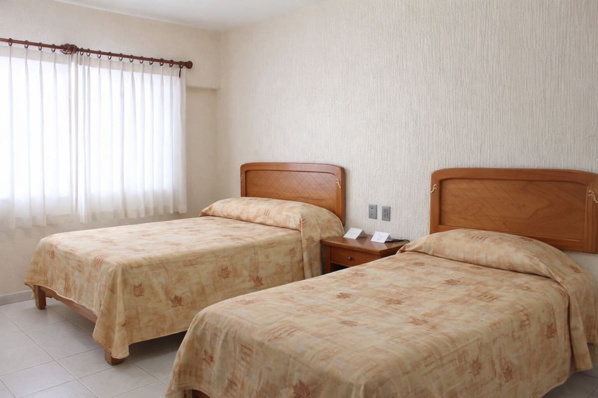 Ett hotellrum med två separata sängar, en sänggavel i trä, ett nattduksbord med en telefon och ett litet bord samt ett fönster med tunna, genomskinliga gardiner.