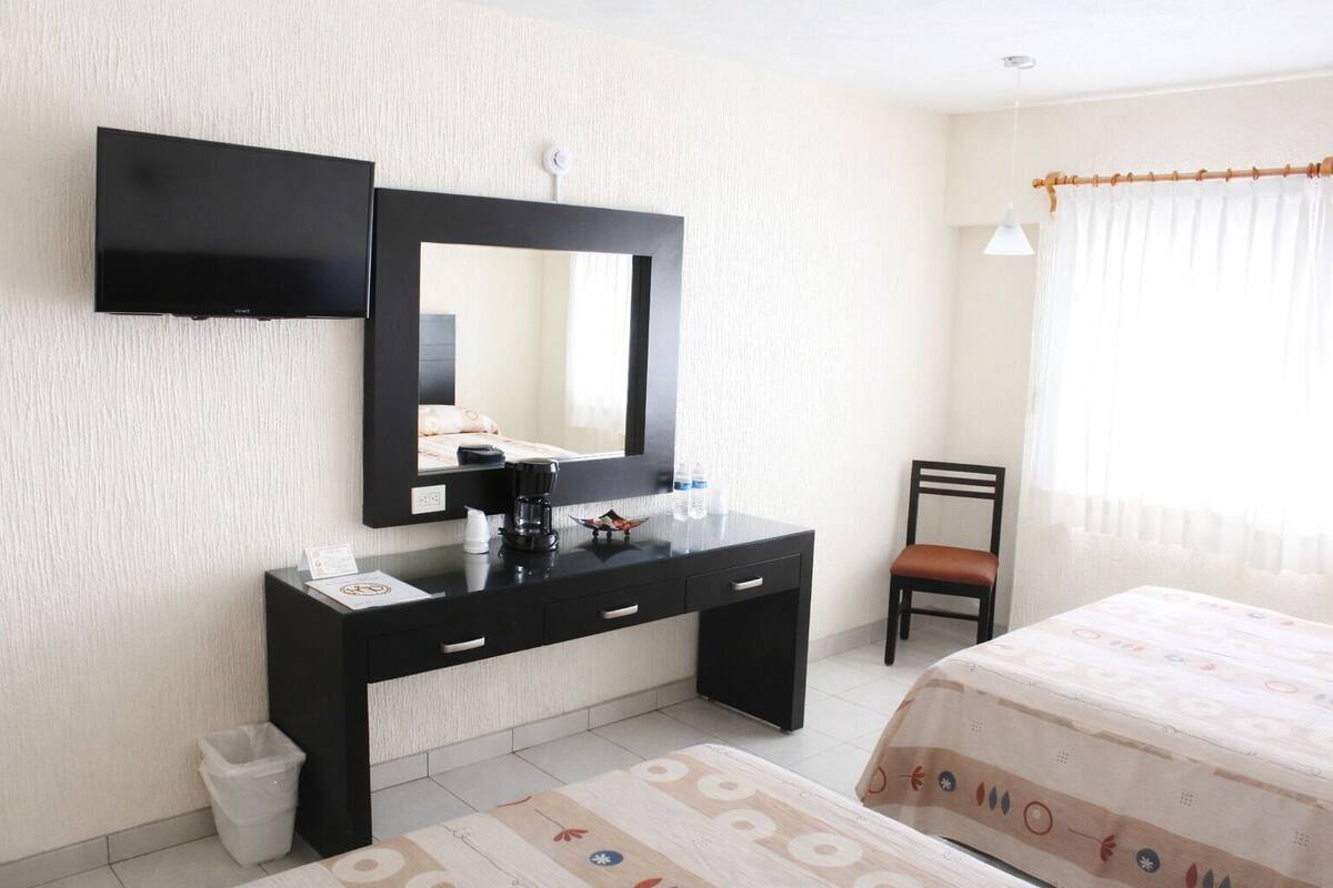 Ett hotellrum med ett svart skrivbord, en stor spegel, en säng med ett mönstrat överkast, en stol och en liten papperskorg.