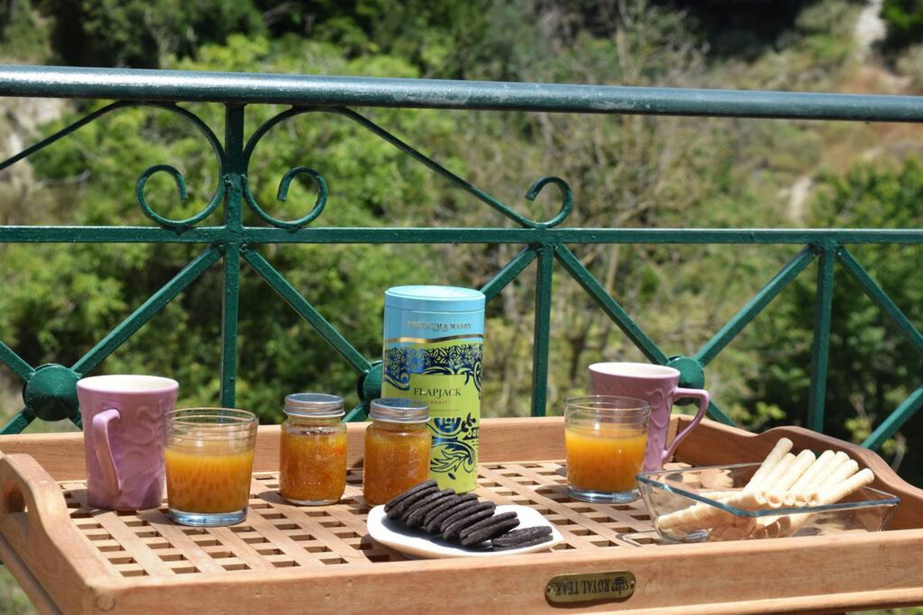En bakke med morgenmadsprodukter, herunder en dåse Flapjack, to kopper, to glas appelsinjuice, en tallerken med småkager og et glas mælk på en balkon med gelænder i grøn farve.