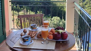 A balcony with a table set for breakfast, including eggs, pastries, and fruit, overlooking a lush landscape.