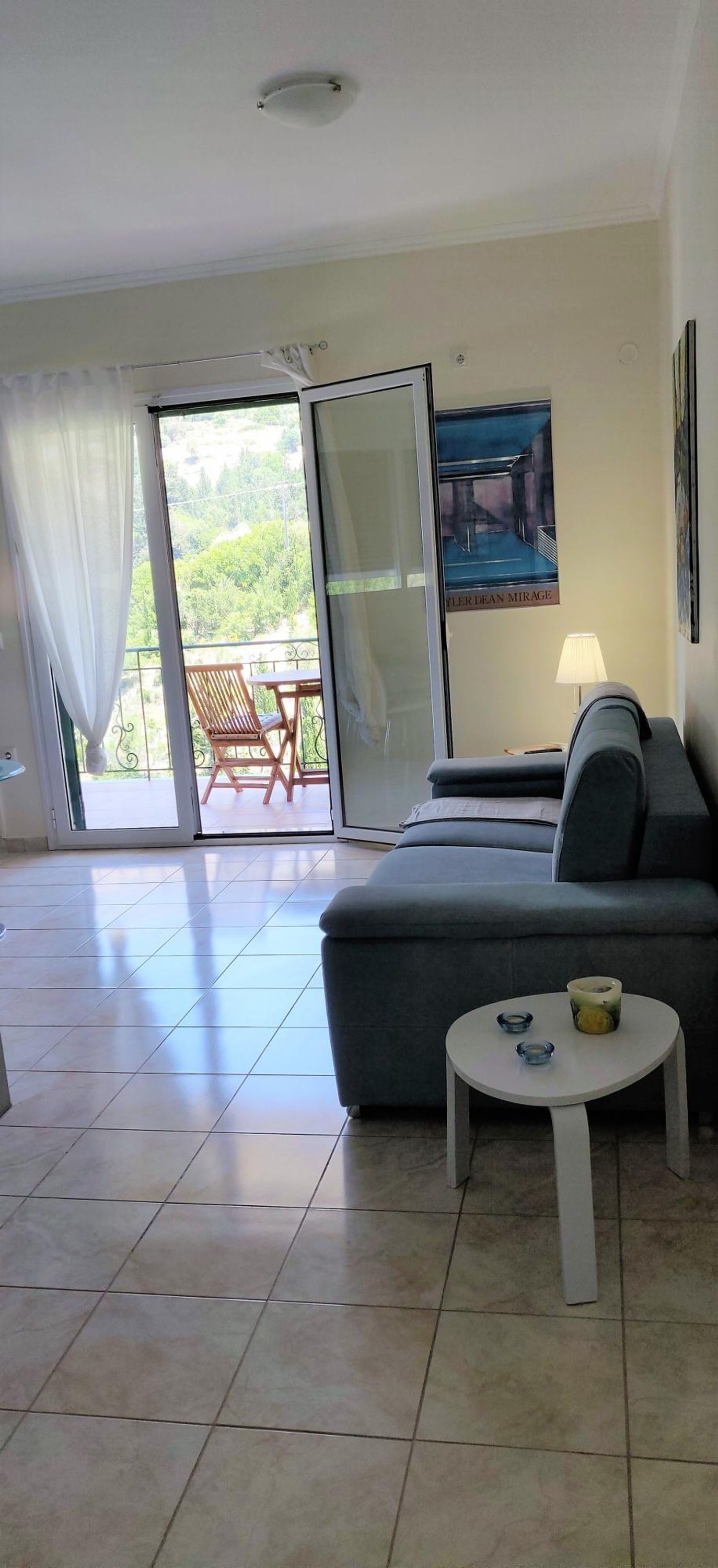 Una sala de estar con un sofá gris, una mesita redonda con una lámpara, y un balcón con una silla y una mesa.