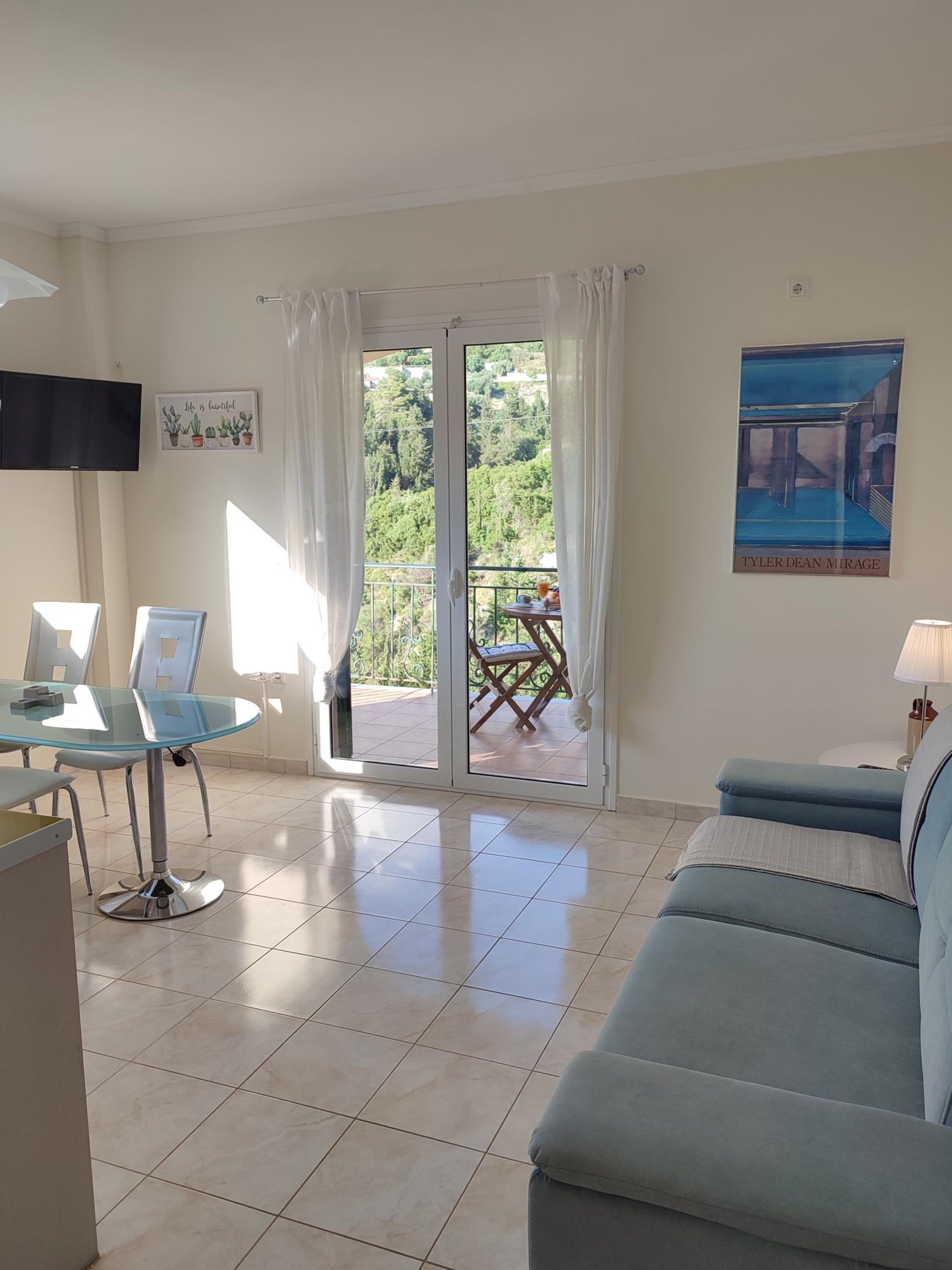 Un salon avec une table en verre, un canapé bleu, un téléviseur fixé au mur et un balcon avec une table et des chaises.