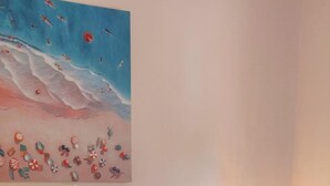 Ein ordentlich bezogenes Bett mit weißen Leinentüchern und einem zusammengerollten Handtuch, ein Nachttisch mit Lampe und ein Gemälde mit Strandmotiv an der Wand.