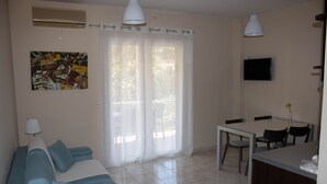 Un salon comprenant un canapé, une table à manger avec des chaises, un téléviseur fixé au mur et une fenêtre avec des rideaux.