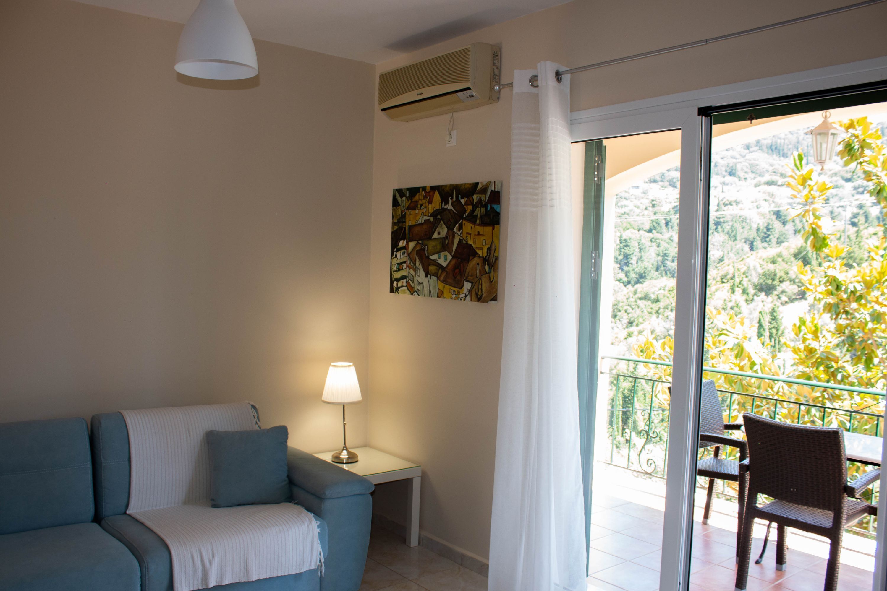 A room with a blue sofa, a small table with a lamp, a painting on the wall, and a balcony with a chair.