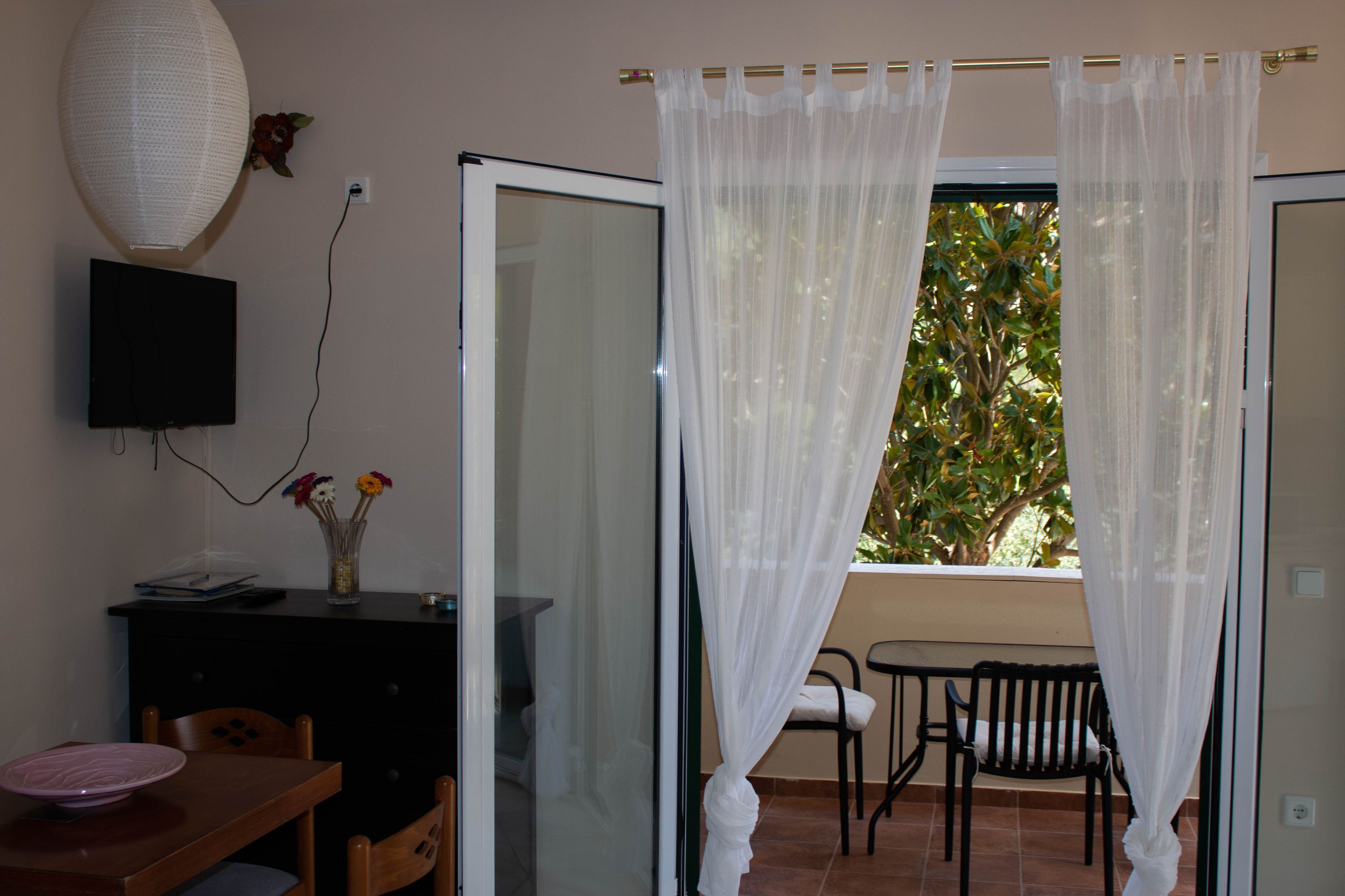 A room with a dining table, chairs, a TV mounted on the wall, a vase with flowers, and a window with sheer curtains.