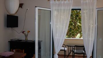 A room with a dining table, chairs, a TV mounted on the wall, a vase with flowers, and a window with sheer curtains.