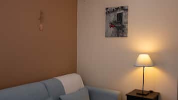 A room with a blue sofa, a small table with a lamp, a wall-mounted picture, and a wall clock.