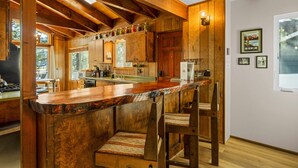 Una cocina rústica con vigas de madera, una zona de bar con taburetes y un ventanal con vistas a árboles.