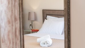 Una cama bien hecha con sábanas blancas, una toalla doblada y una lámpara de noche en una mesita.