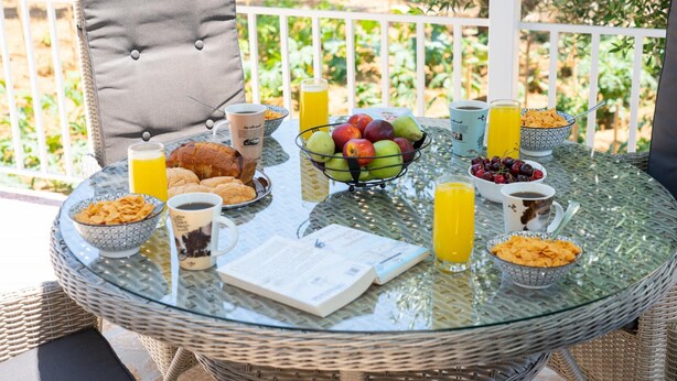 Una mesa de vidrio con un sillón de mimbre, mostrando tazones de cereal, frutas y bebidas para el desayuno.