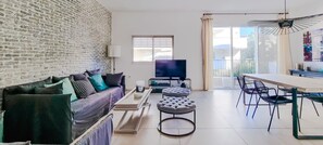 A modern living room with a brick wall, a sofa with cushions, a TV, a coffee table, an ottoman, and a dining area with chairs. - Hovevei Tsiyon Magnolia (Tel Aviv-Yafo)