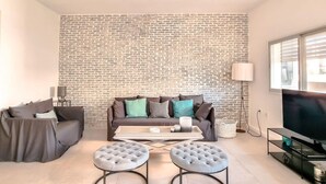 A living room with a brick wall, a sofa, two ottomans, a coffee table, a TV, and a lamp. - Hovevei Tsiyon Magnolia (Tel Aviv-Yafo)