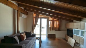 A cozy living area with a wooden ceiling, a sofa, a dining table, and a kitchen area with cabinets and a TV. - Samaras Apartments - Sea View Attic (Kamena Vourla)