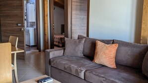 A modern living room with a grey sofa, wooden ceiling beams, and a wooden door leading to a kitchen. - Samaras Apartments - Sea View Attic (Kamena Vourla)