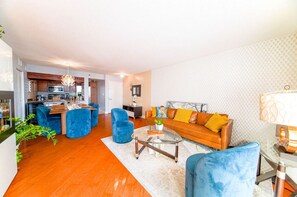 Een moderne woonkamer met een bank, blauwe stoelen, een glazen salontafel en een keuken op de achtergrond.