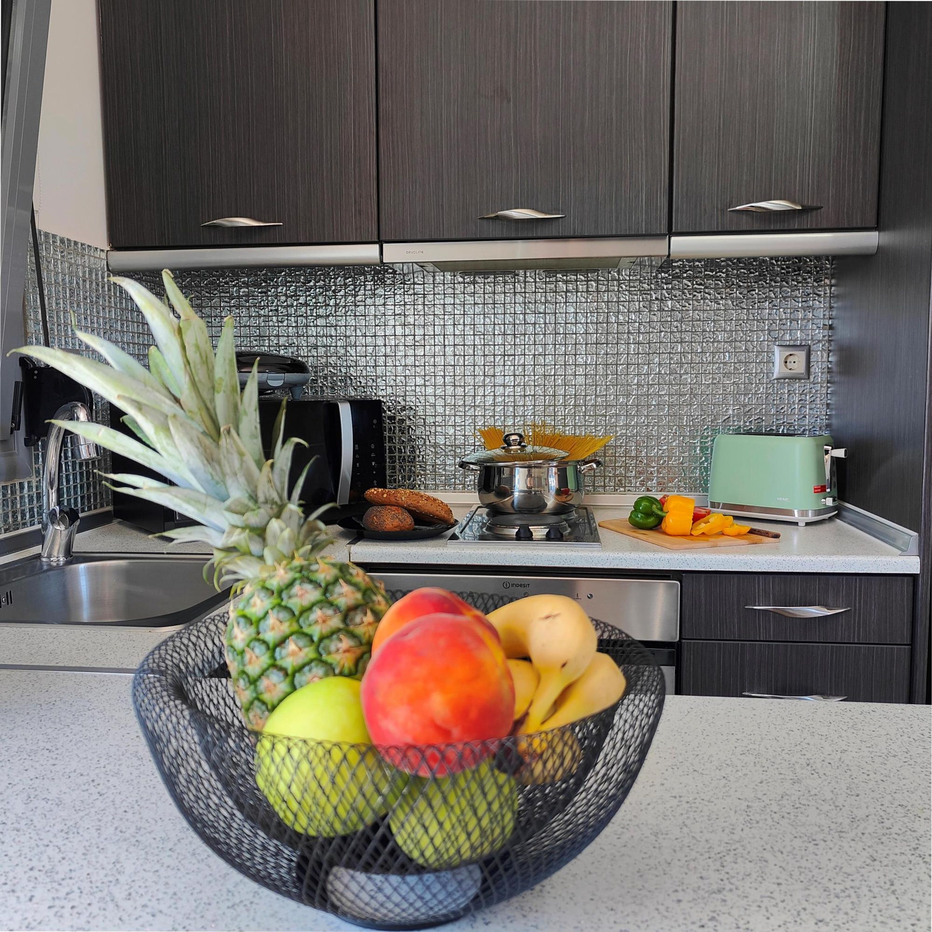 Una cocina con armarios oscuros, un salpicadero de azulejos, un frutero con piña, mango y plátano, un tostador y un hervidor.