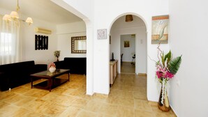 Zona d'estar àmplia amb un sofà de color fosc, una taula de centre amb superfície de vidre i un ramader decoratiu amb flors. Una arcada condueix a la cuina, que disposa d'una petita illa i una làmpada penjant.