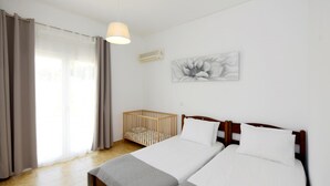 Habitació d'hotel amb dos llits, una bressol, una finestra gran amb cortines, aire condicionat fixat a la paret i una làmpada al sostre.