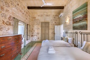 A room with stone walls, a wooden dresser, two beds, a ceiling fan, and a painting on the wall. - Kikka - Xaghra Holiday Home (Ix-Xagħra)