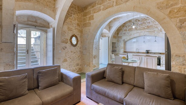 A spacious living area with stone walls and arches, a dining area with a kitchen visible through an archway, and a sofa with cushions. - Kikka - Xaghra Holiday Home (Ix-Xagħra)