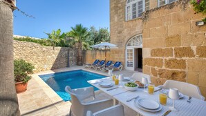 A poolside dining area with a table set for breakfast, lounge chairs, and a stone wall. - Kikka - Xaghra Holiday Home (Ix-Xagħra)