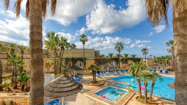 Un espace piscine aménagé dans un complexe hôtelier, avec des palmiers, des chaises longues et un ciel d’un bleu limpide.