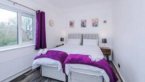 Un dormitorio con un ventanal grande, una cama con sábanas blancas y mantas moradas, una mesita de noche con una lámpara y un reloj de pared.