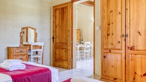 A hotel room with two beds, a wooden wardrobe, a desk with a chair, and a dining area visible through an open door. - Villagg tal-Fanal - 1BDR (L-Għasri)
