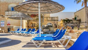 A pool area with blue lounge chairs and thatched umbrellas, surrounded by stone buildings and greenery. - Villagg tal-Fanal - 1BDR (L-Għasri)