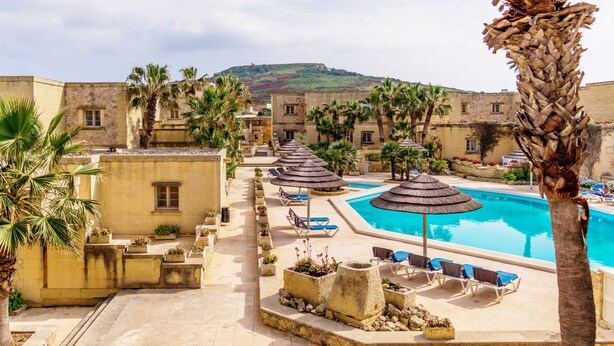 A resort pool area with lounge chairs and umbrellas, surrounded by palm trees and stone buildings. - Villagg tal-Fanal - 1BDR (L-Għasri)