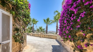 A narrow stone pathway flanked by walls covered with purple bougainvillea, leading to a view of palm trees and distant hills. - Gharilma IV – Santa Lucija Holiday Home (Ta' Kerċem)