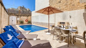 A poolside area with lounge chairs, a table with chairs, and a grill. - Gianni - Xewkija Holiday Home (Ix-Xewkija)