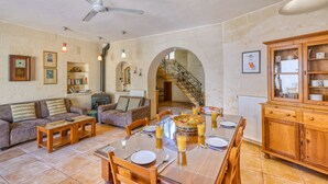 A spacious living area with a dining table set for two, a fireplace, and a staircase leading to an upper level. - Gianni - Xewkija Holiday Home (Ix-Xewkija)