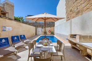A poolside area with a table set for dining, lounge chairs, and an umbrella. - Gianni - Xewkija Holiday Home (Ix-Xewkija)