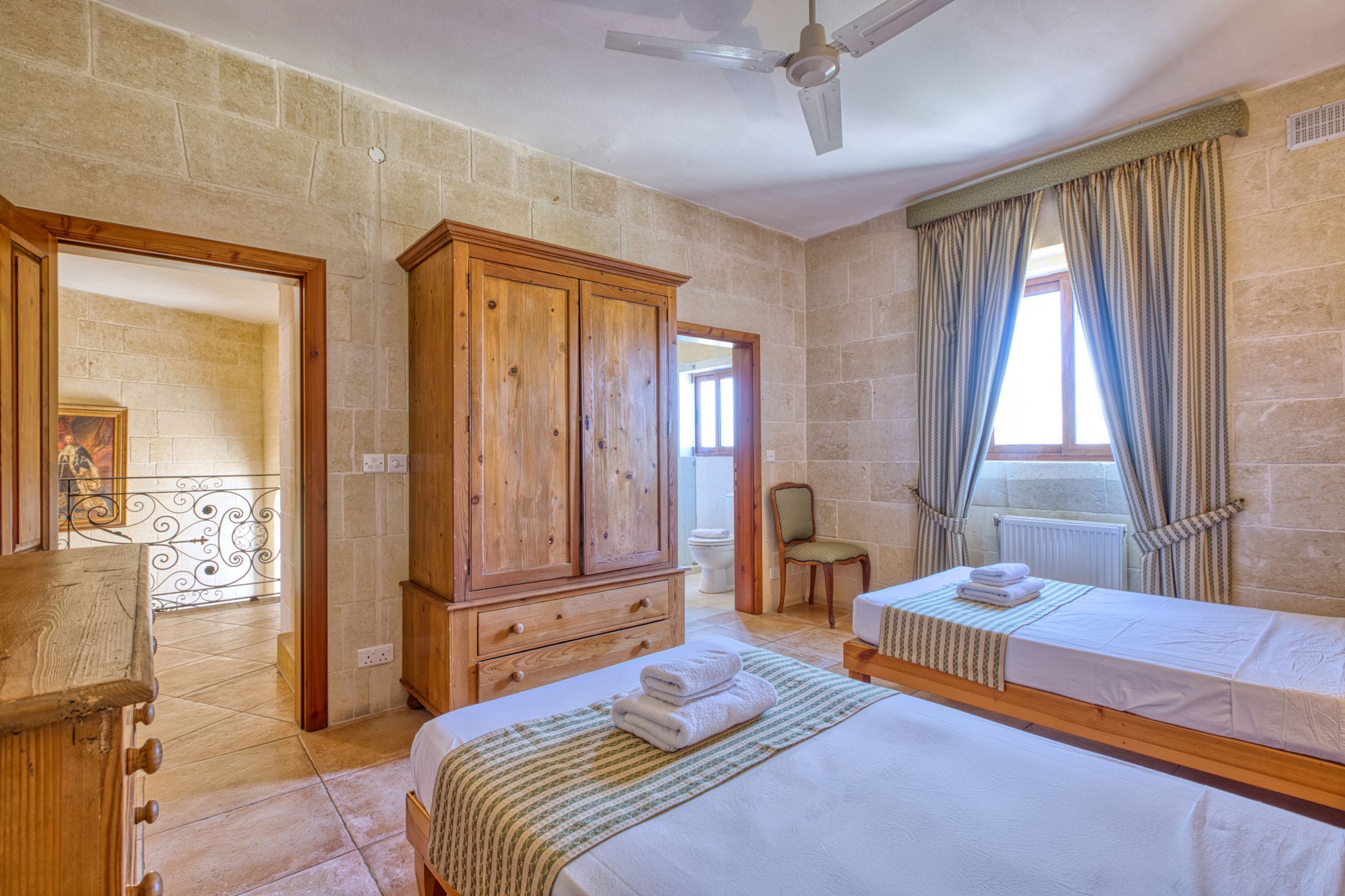 A hotel room with a large wardrobe, a bed with a striped bedspread, a bedside table, and a window with curtains.