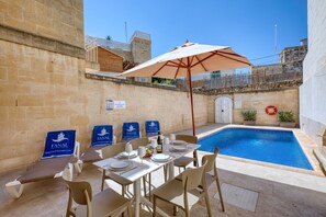 A poolside area with a table set for dining, a pool, and a stone wall. - Gianni - Xewkija Holiday Home (Ix-Xewkija)
