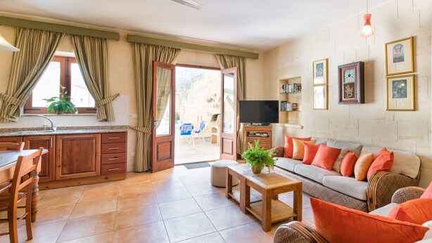 A spacious living area with a sofa, coffee table, and dining table. There is a television, a bookshelf, and a window with curtains. - Gianni - Xewkija Holiday Home (Ix-Xewkija)