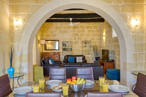 A dining area with a stone wall, a ceiling fan, and a table set for breakfast with plates, glasses, and a fruit bowl.