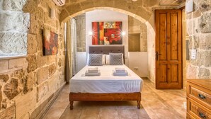 Une chambre avec un mur en pierre, un lit en bois, un ventilateur de plafond et une porte.