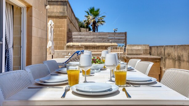 Une table mise pour le petit-déjeuner, avec des chaises blanches, des assiettes, des couverts et des verres de jus d’orange.