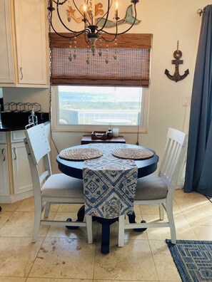 Ein Essbereich mit einem runden Tisch, zwei weißen Stühlen, einer gemusterten Tischdecke, einer Chandelüstere, Blick durch ein Fenster mit Jalousien und einem kleinen Tisch mit Kerze und Flasche.