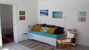 A room with a white sofa, a small side table, a chair, a standing fan, and a cart with items. - Amberi, Tinos in Panormos (Panormos)