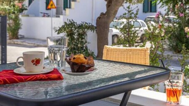 Een glazen tafel met een kop, een drankje en een muffin op een balkon, met een boom erboven.