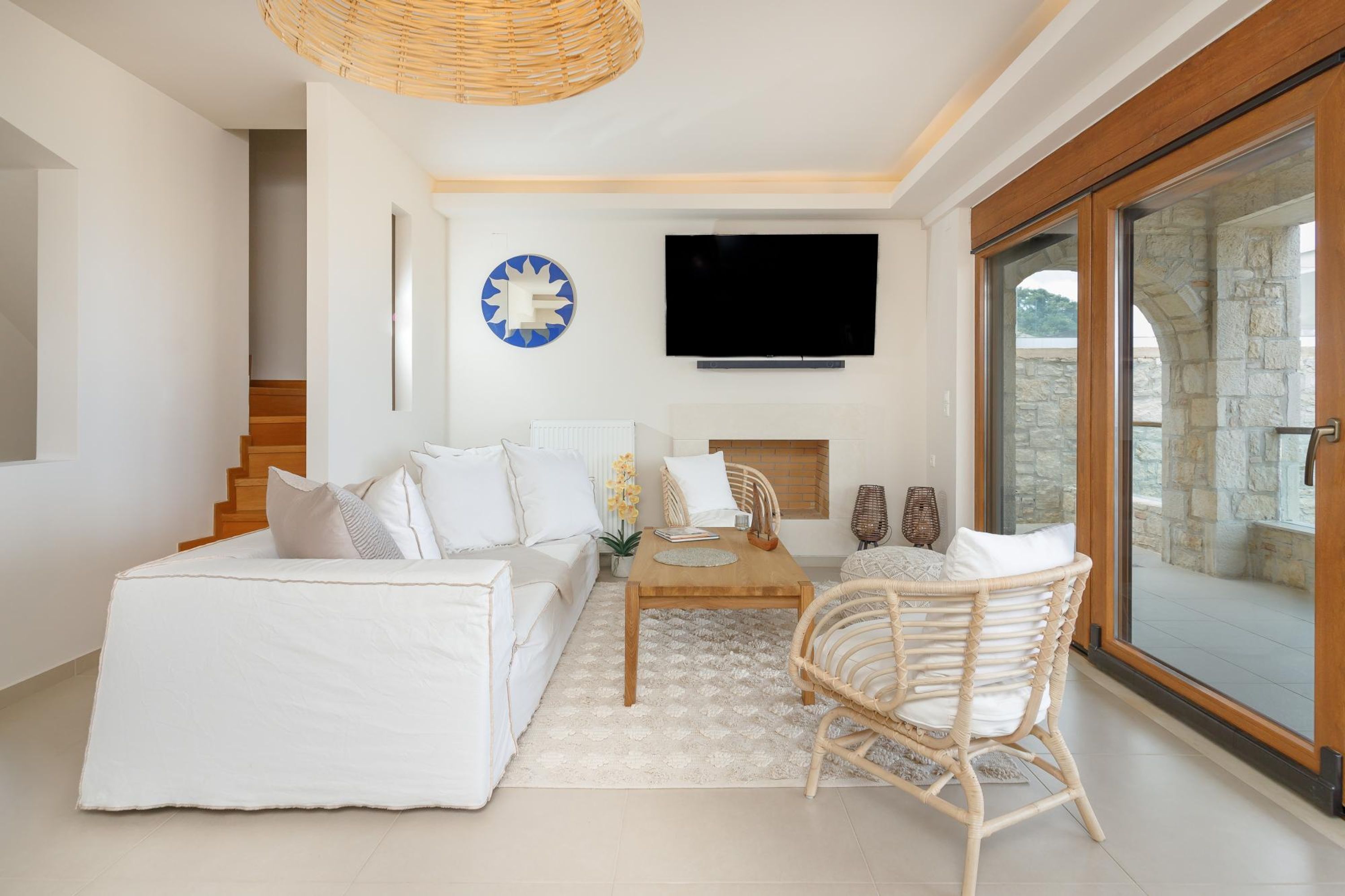 A modern living room with a white sofa, a wooden coffee table, and a wicker chair. A flat-screen TV is mounted on the wall above a fireplace.